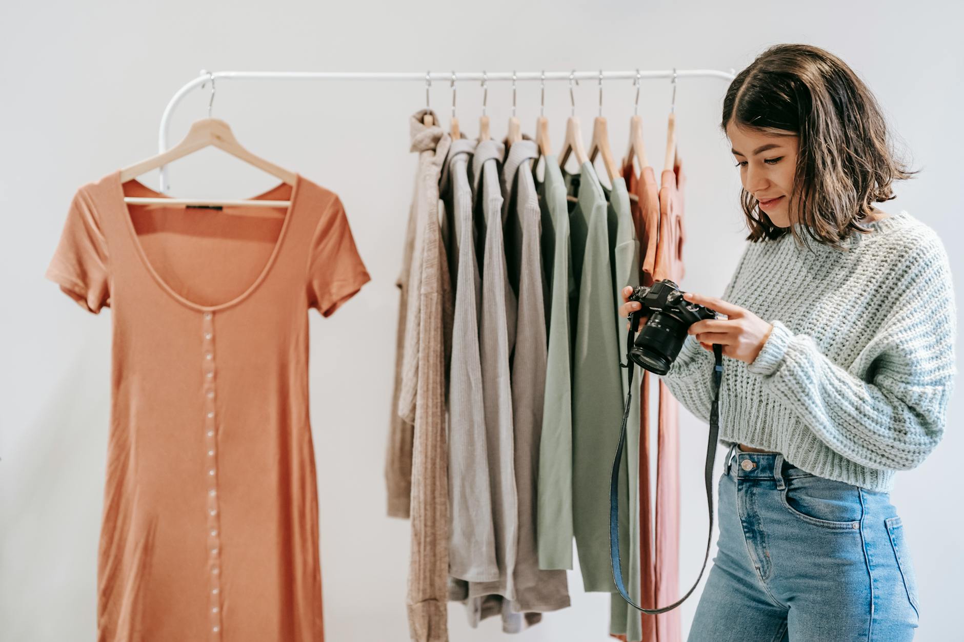 Young smiling Latin American woman with professional photo camera against rack with assorted apparel indoors
