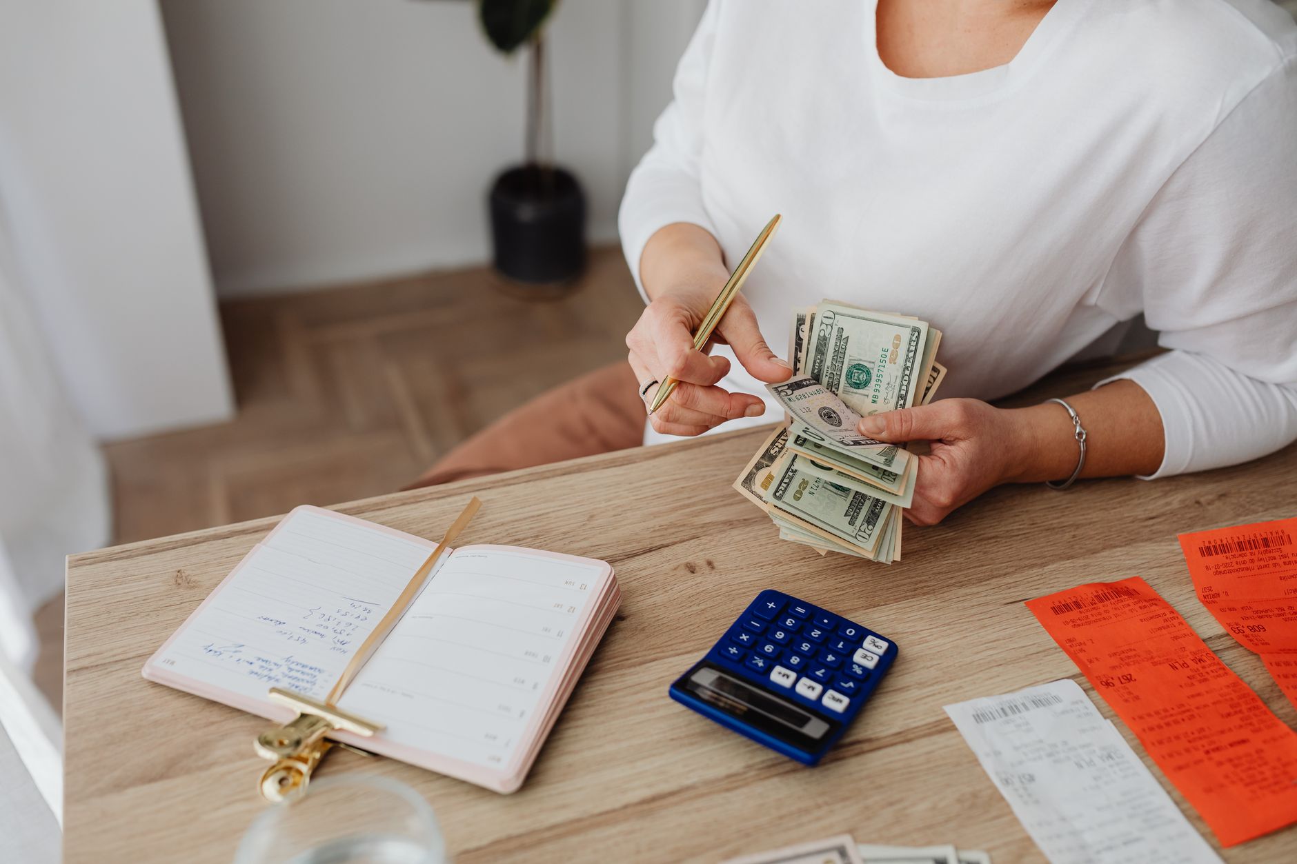 A person counting bank notes and coins while writing a business budget at a kitchen table