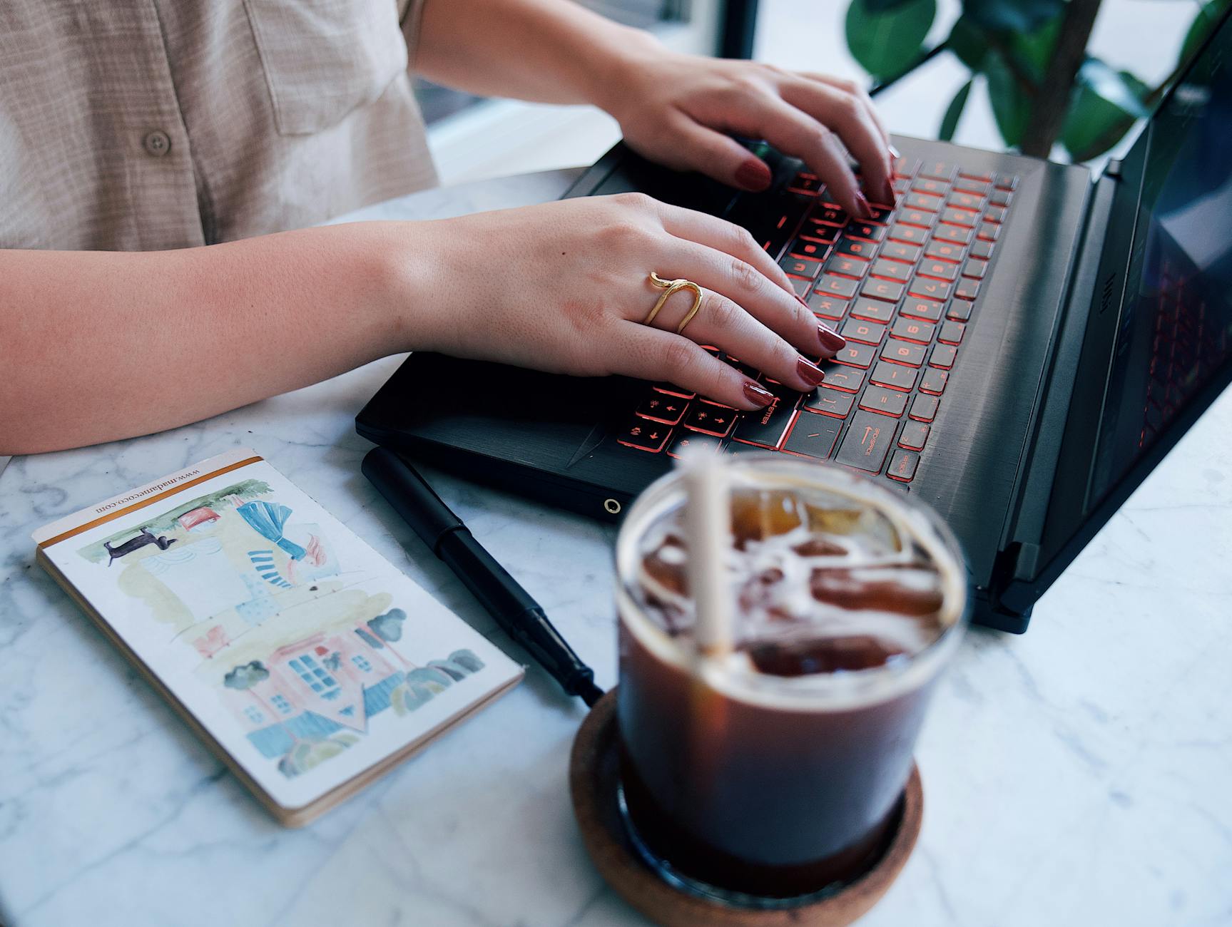 A woman typing on a laptop at a marble table with an iced coffee and a notepad.