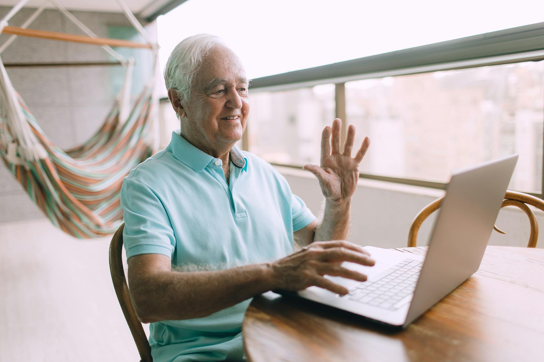 Elderly man engaging in a video call using a laptop from his home, sitting comfortably near a hammock.