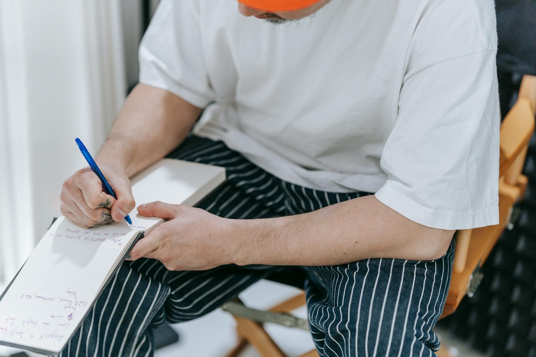 A man sits indoors writing notes with a pen in a notepad. Casual setting.