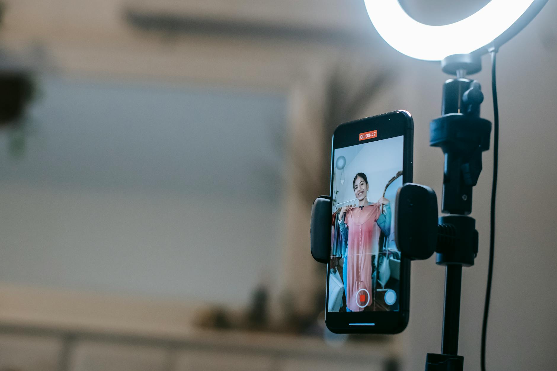 Anonymous female blogger standing in room and shooting vlog on smartphone with ring lamp while demonstrating pink dress in hands