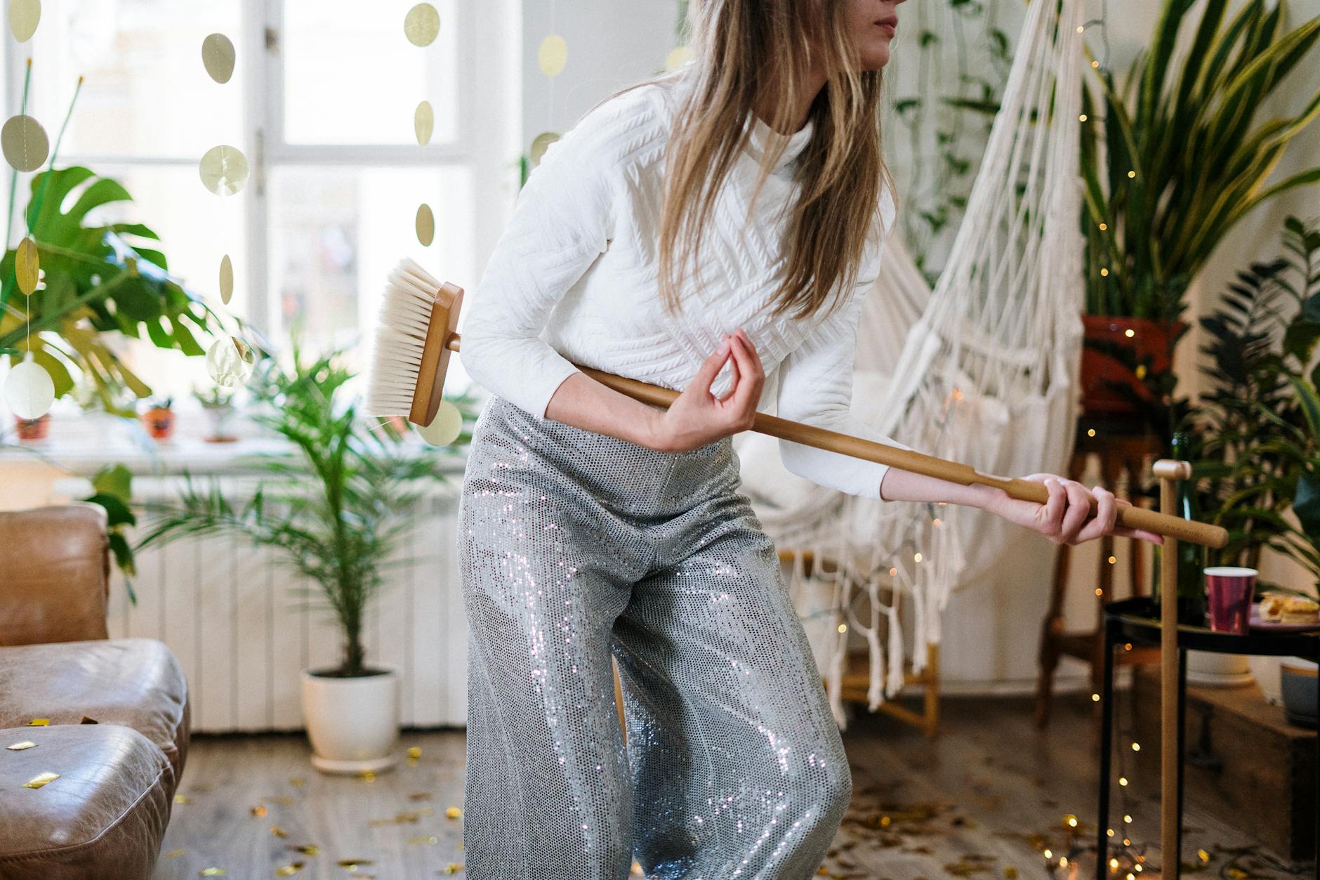 Energetic woman in sparkling pants mimicking guitar play with broom amidst lively indoor setting.