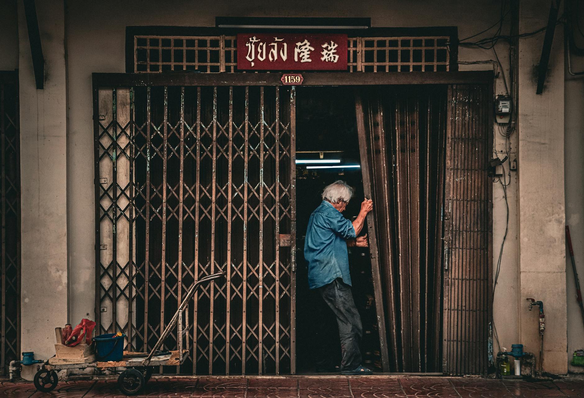 Elderly man closing a metal gate of an urban shop in Asia with traditional signage.
