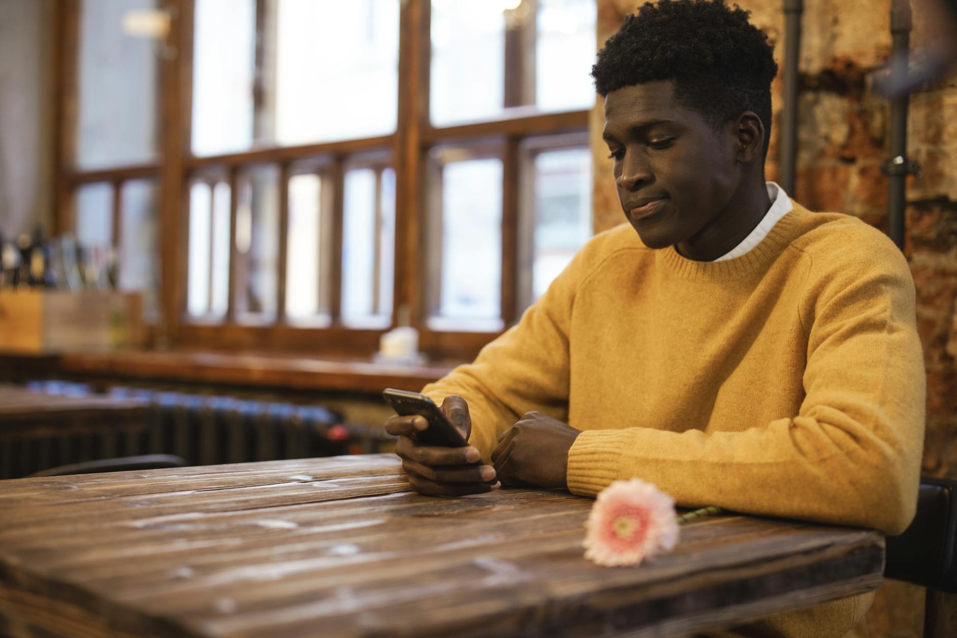 A man in a café using a smartphone, sitting at a wooden table with a flower, creating a warm and inviting atmosphere.