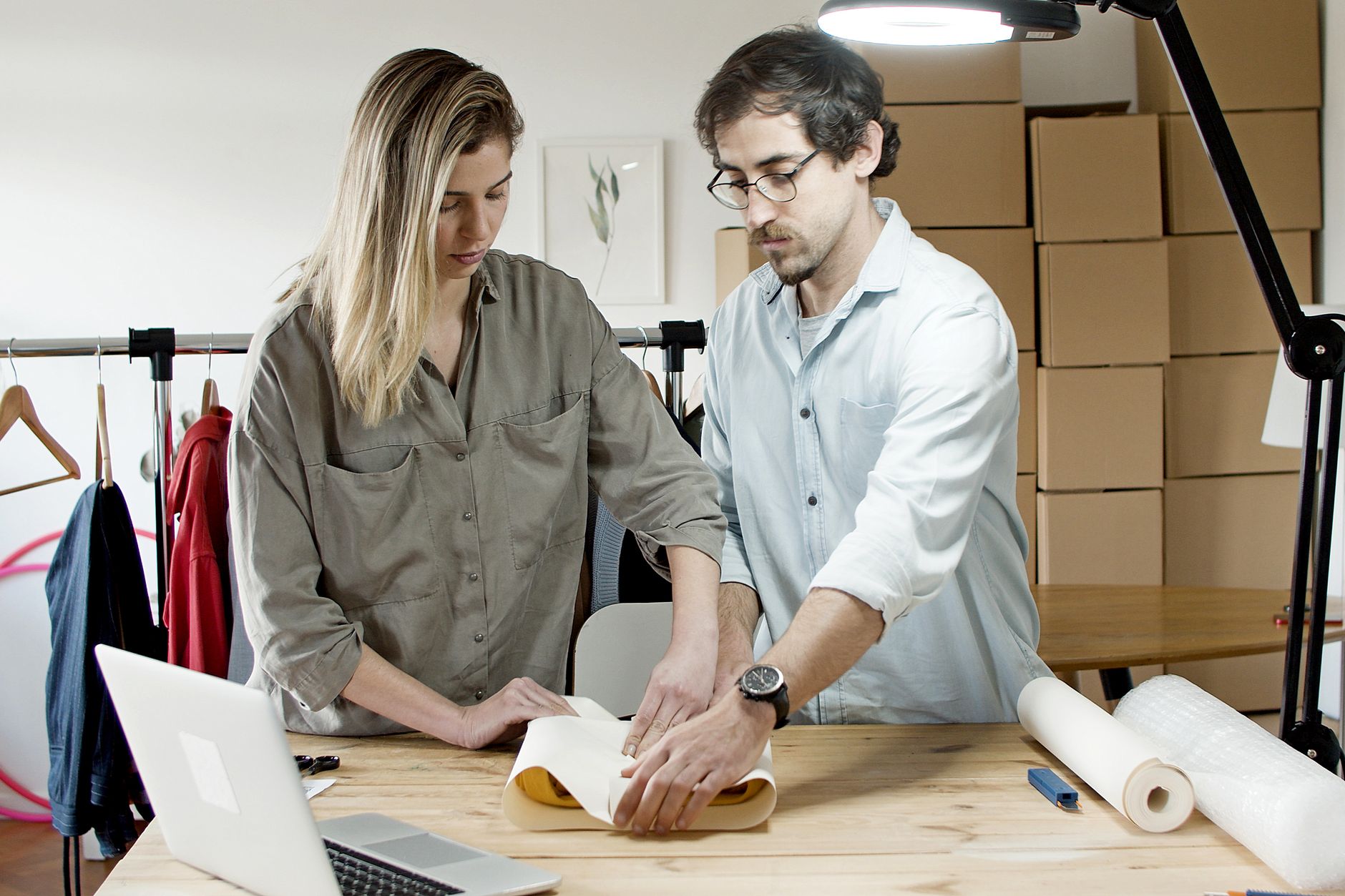 An artist carefully rolls a signed print into a shipping tube at a worktable stacked with packaging, fulfilling an online order.