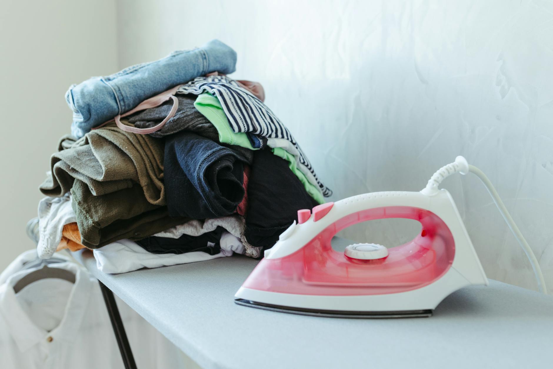 Tidy stack of clothes ready to be ironed with an iron on an ironing board.
