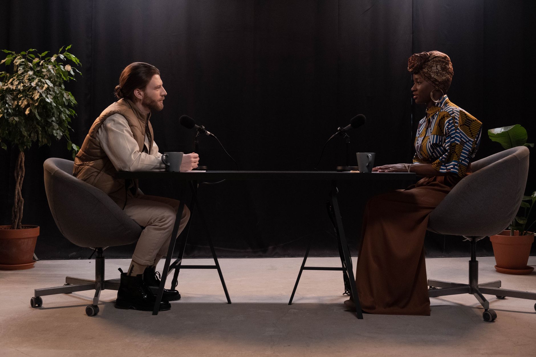 Two people sit across a table recording a podcast interview, each speaking into their own microphone with headphones on.